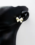 Gold bow-shaped earring on a black mannequin ear against a white background
