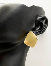 Soft square matte gold earrings resting on textured fabric.